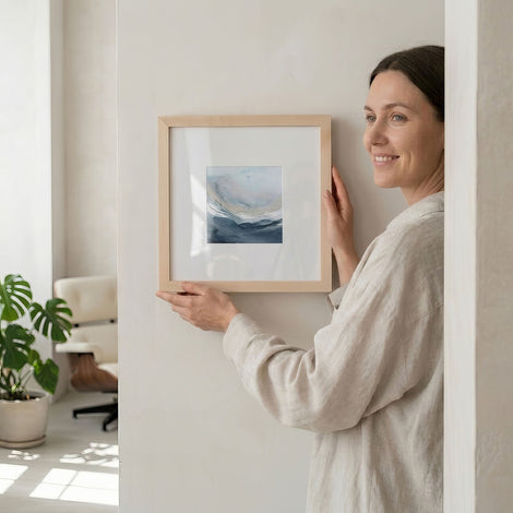Woman hanging Nourishing Waters No. 2 — 14x14 coastal abstract fine art print with white mat in natural maple frame by Julia Contacessi in her home.